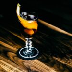Copa de vino de color ámbar anaranjado junto a racimos de uva blanca sobre una mesa de madera